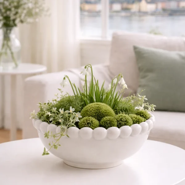 „Weiße Keramikschale mit Perlenrand, dezent frühlingshaft bepflanzt mit Grünpflanzen und Moos, ruhig inszeniert im hellen Wohnzimmer im nordischen Sti