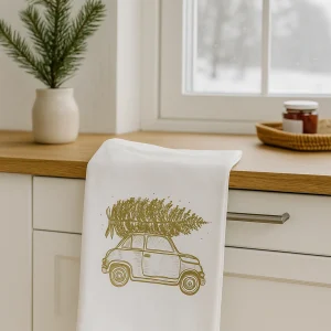 Weißes Geschirrtuch mit goldener Auto-Illustration und Tannenbaum, locker über einer Schubladenleiste in einer hellen skandinavischen Winterküche mit Tannenzweig und Blick auf verschneite Landschaft