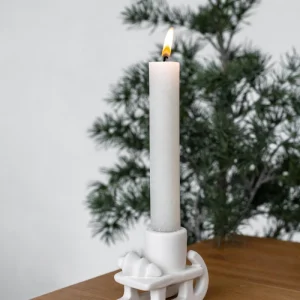 Weißer, nordischer Kerzenhalter in Schlittenform mit liegendem Tannenbaumdetail und brennender Stabkerze, dekoriert mit Tannenzweig auf hellem Leinenuntergrund