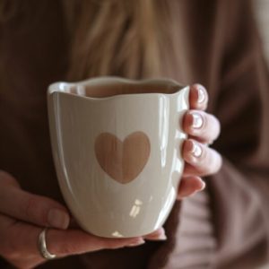 Beige Steinzeug-Becher mit handgemaltem Herzmotiv, in Händen gehalten, warme gemütliche Stimmung