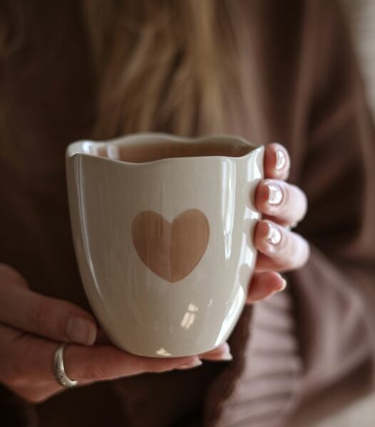 Beige Steinzeug-Becher mit handgemaltem Herzmotiv, in Händen gehalten, warme gemütliche Stimmung