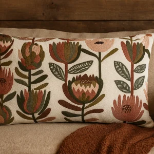 Dekokissen mit floraler Stickerei in warmen Naturtönen auf einem beigen Sofa vor einer rustikalen Holzwand. Erdige Farben wie Salbeigrün, Terrakotta und Rosé bilden ein botanisches Muster im nordischen Stil
