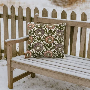 Besticktes quadratisches Dekokissen mit floralen Mustern in Olivgrün und Terracotta auf weißem Stoff, liegend auf einer Holzbank vor einem dänischen Gartenzaun mit leichtem Frost.