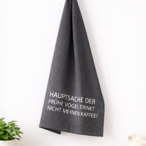 Graues Geschirrtuch mit Aufdruck ‚Hauptsache der frühe Vogel trinkt nicht meinen Kaffee!‘ an schwarzem Haken in heller Scandi-Küche